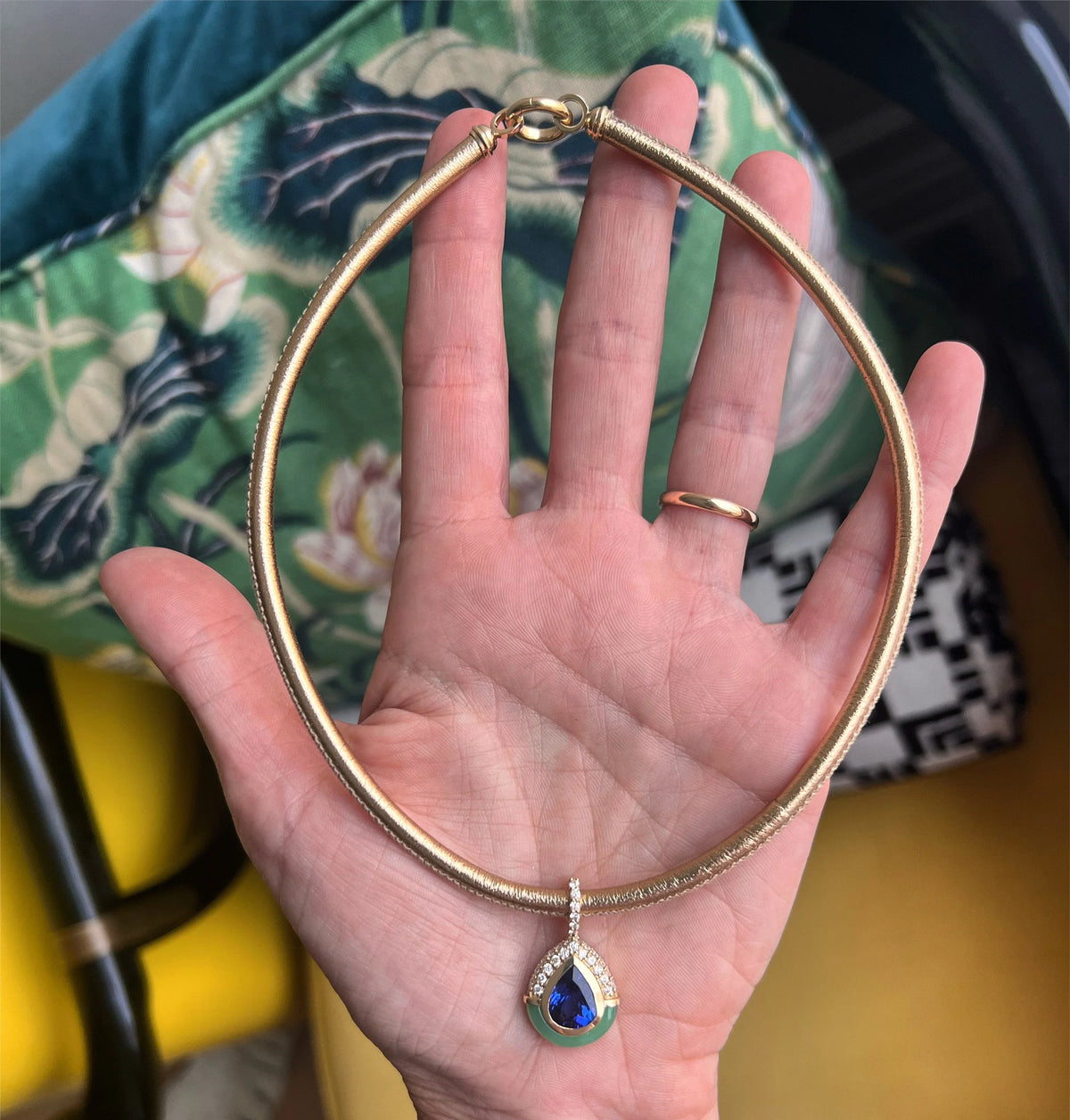 A gold snake chain necklace with a teardrop-shaped pendant featuring a blue and green stone, held in a person's hand.