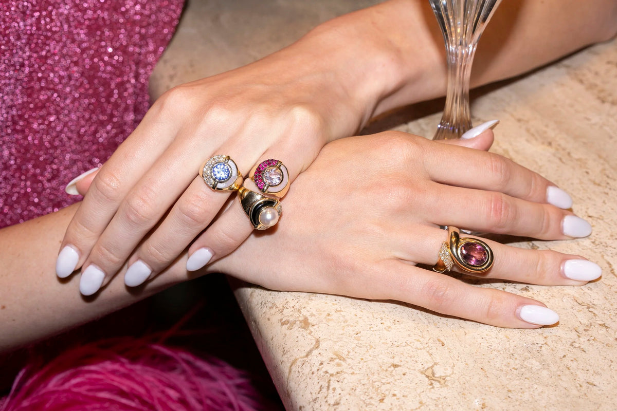 Close-up of a person's hands resting on a stone surface, wearing three gold rings with pink and clear gemstones. A sparkling magenta dress is visible on the left, and part of a champagne flute stands on the right.