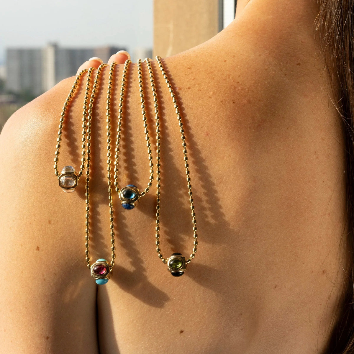 A close-up lifestyle shot of a woman's bare back and shoulder, wearing three gold beaded chains draped over her upper back, each ending in a small glass bead with different colored accents.