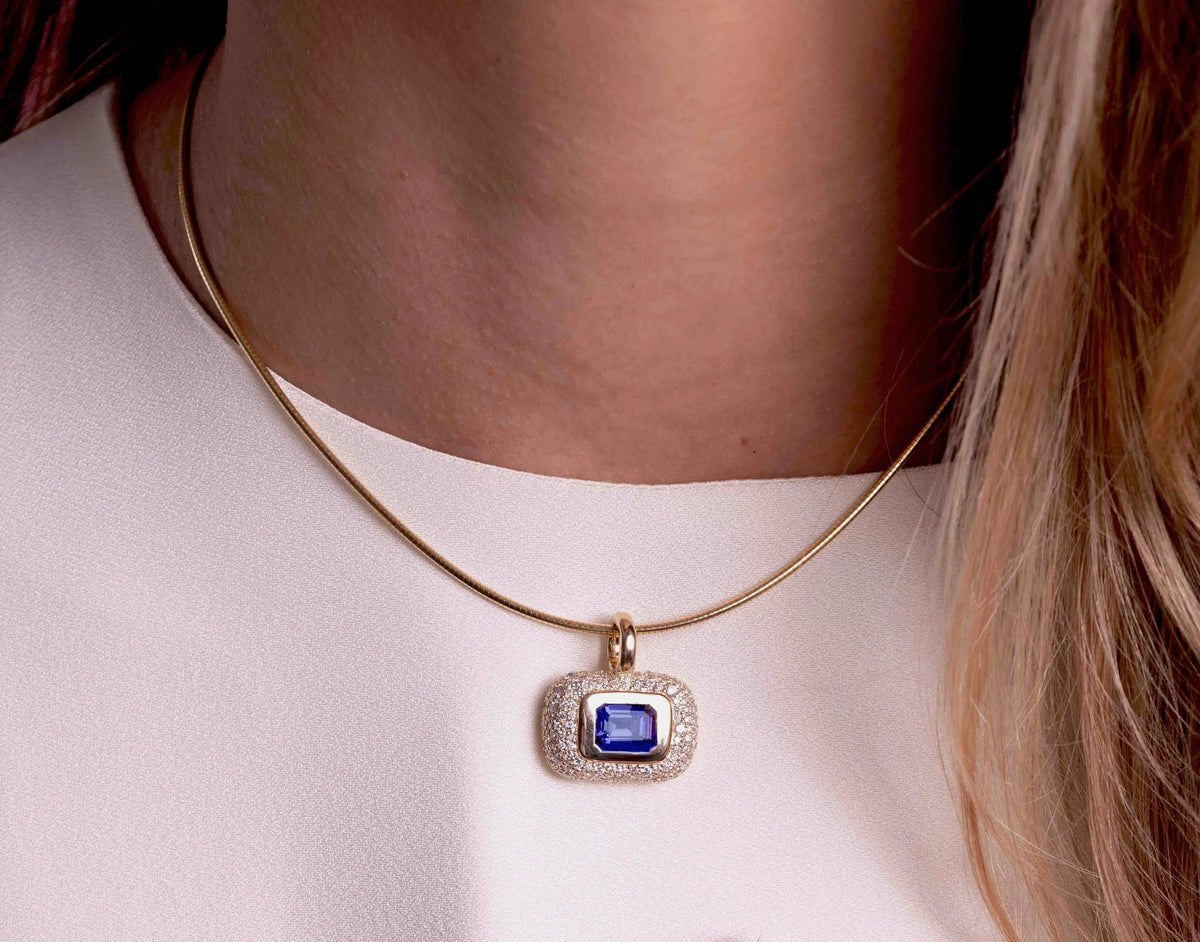 A close-up of a model wearing a delicate gold necklace with a rectangular pendant featuring a deep blue gemstone surrounded by a halo of small clear stones.