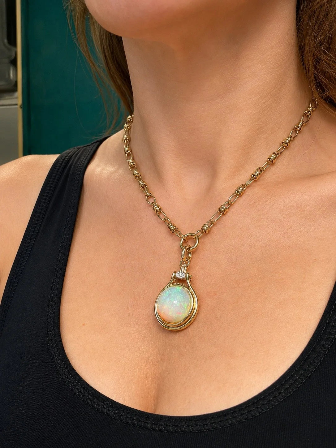 A gold chain necklace with a teardrop-shaped iridescent white opal pendant, worn by a model in a black tank top.