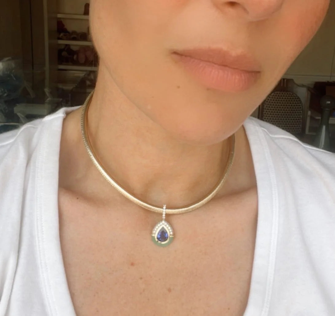 A close-up of a person wearing a delicate gold snake chain choker necklace with a teardrop-shaped pendant featuring a blue and green stone set in silver.