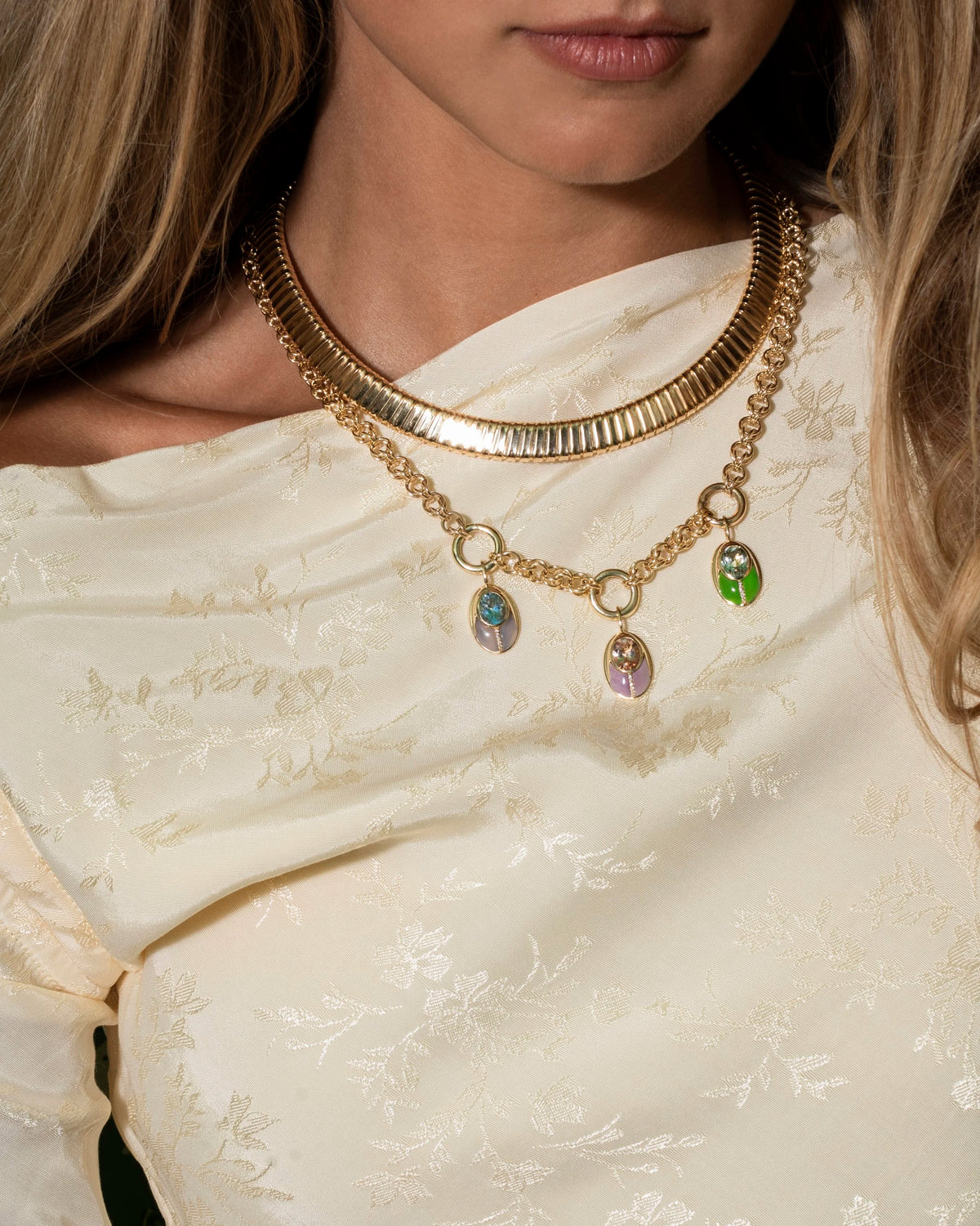 A close-up of a model wearing a layered gold necklace set, featuring a wide ribbed collar choker and a chain necklace with three dangling iridescent oval charms, styled with a cream embroidered top.