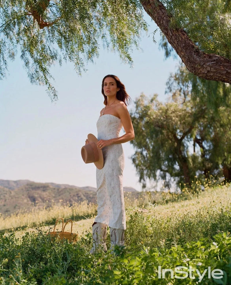 A woman in a white strapless dress holding a brown hat stands in a grassy outdoor field with trees and a mountain in the background on a sunny day.