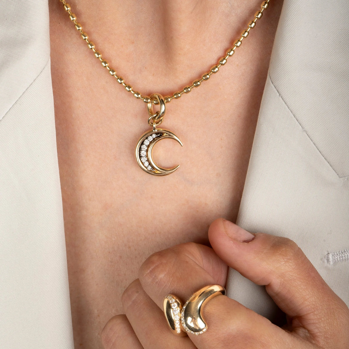 A close-up of a person wearing a gold crescent moon pendant necklace with small clear stones, paired with a matching gold ring on their finger, set against a light beige blazer.