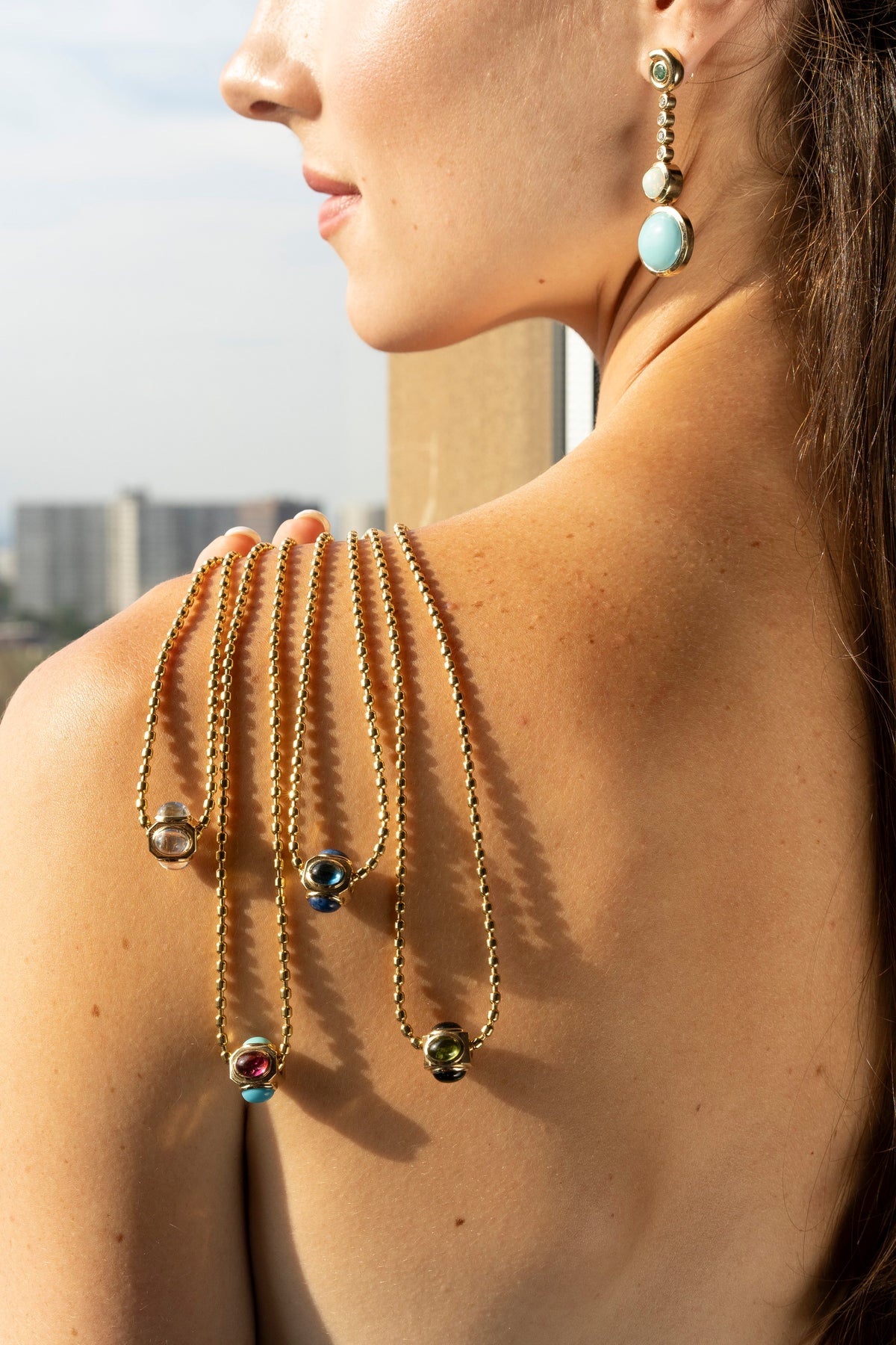 A woman shown from the back wears a set of gold chain necklaces draped over her shoulders, each adorned with colorful glass beads, alongside matching drop earrings with light blue and gold beads.