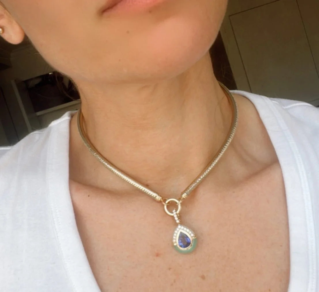A close-up of a person wearing a gold snake chain necklace with a teardrop-shaped pendant featuring a blue and green stone.