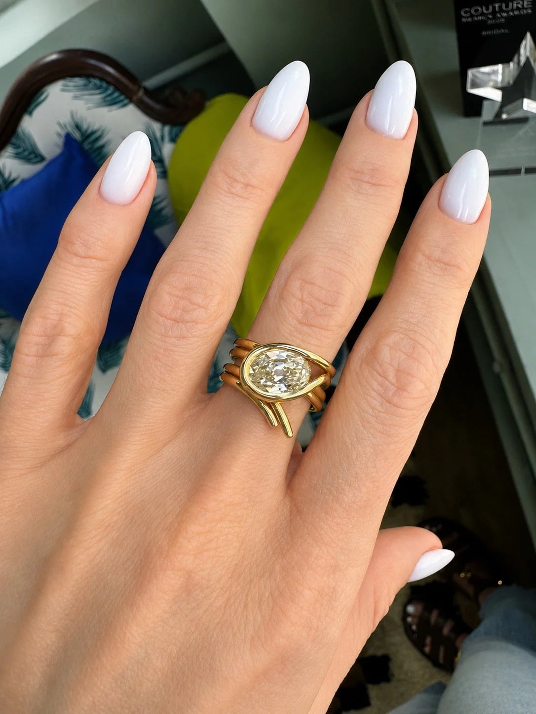 A close-up of a hand with white manicured nails wearing a gold ring with a large oval clear gemstone and smaller side stones, set against an indoor background.