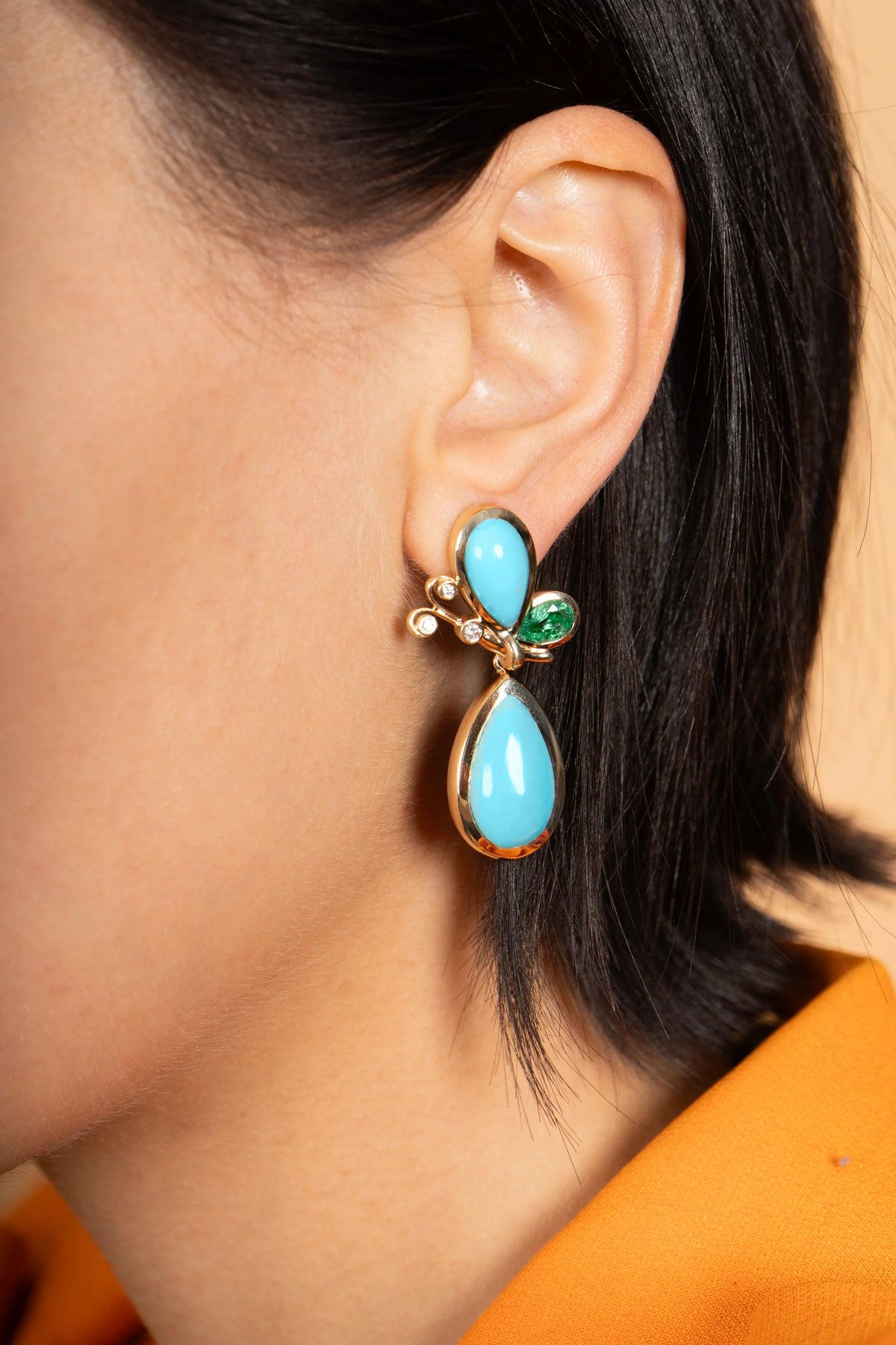 A close-up of a model's ear wearing a gold-toned drop earring with a light blue teardrop stone and small green and blue accent stones.