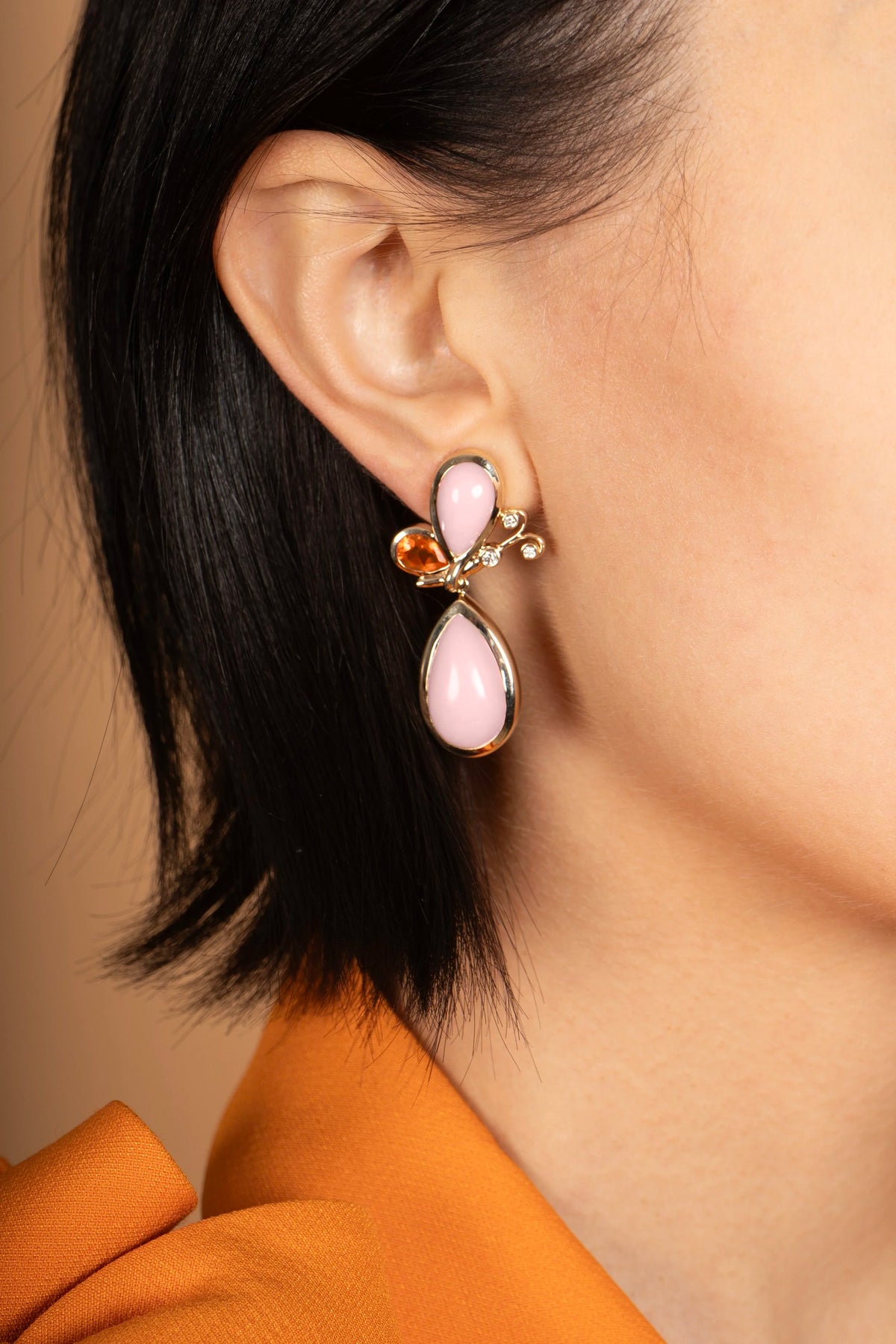 A close-up of a model's ear wearing a dangling earring with a light pink teardrop-shaped stone, a small orange stone, and gold-toned metal details.
