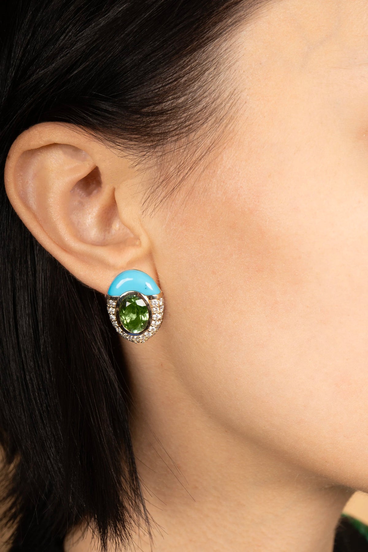 A close-up of a model's ear wearing a unique stud earring featuring a bright turquoise blue crescent-shaped top, a central green oval gemstone surrounded by small diamonds, and a silver-toned base.