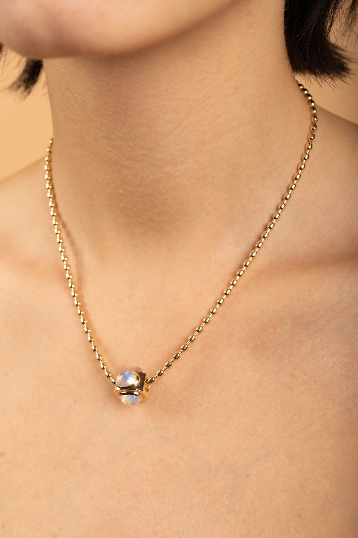 A close-up of a model's neck and upper chest wearing a delicate gold chain necklace with a small gold bow pendant holding a clear, iridescent glass bead.