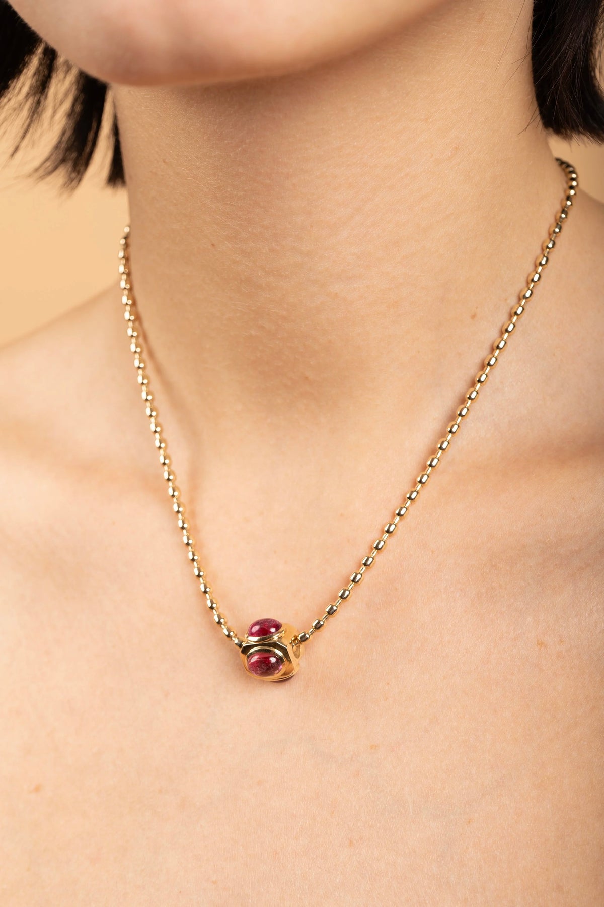 A close-up of a model wearing a delicate gold chain necklace with a small gold and red bead pendant.