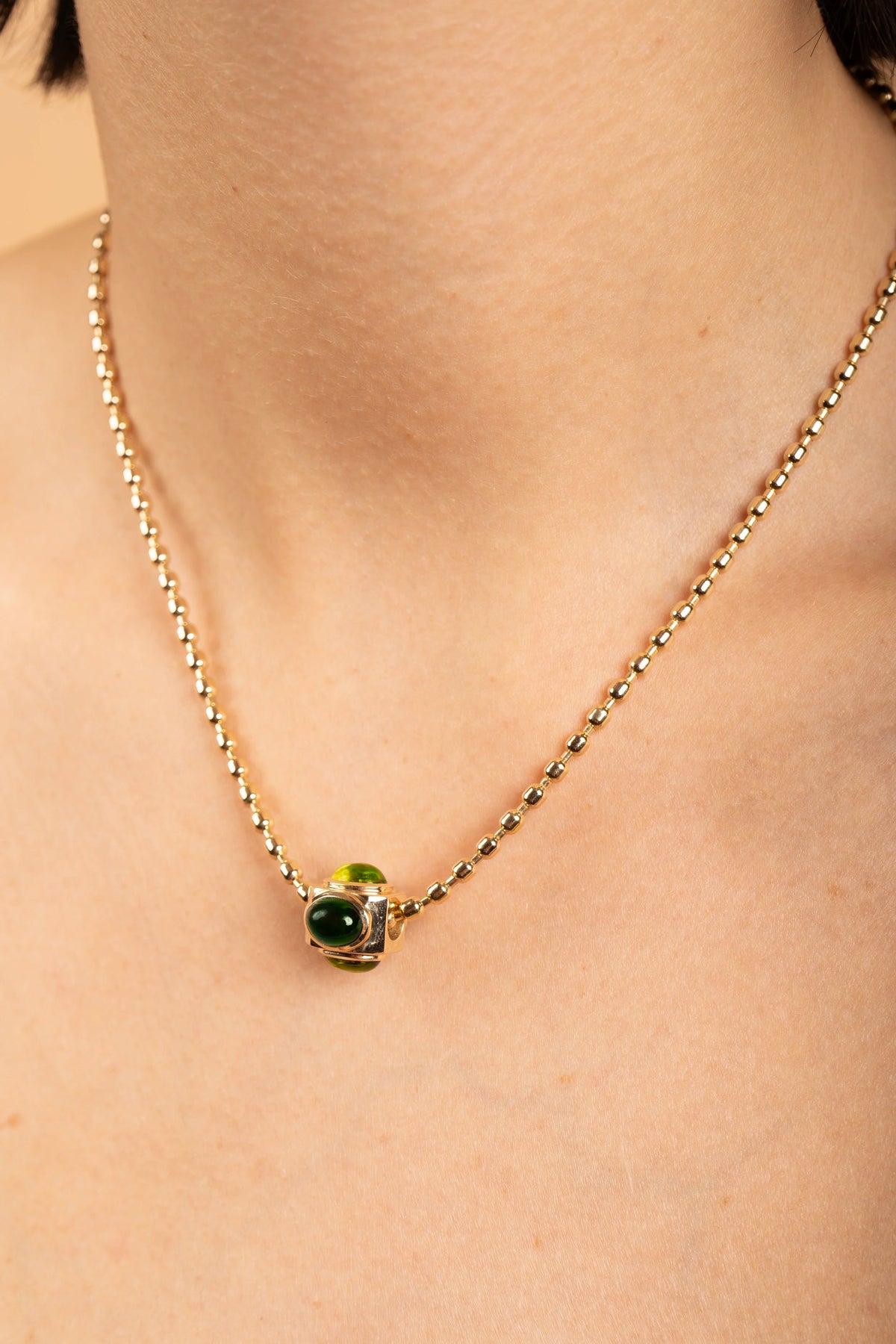 A close-up of a person's neck and chest wearing a delicate gold chain necklace with a small pendant featuring a green oval stone and a light green accent.