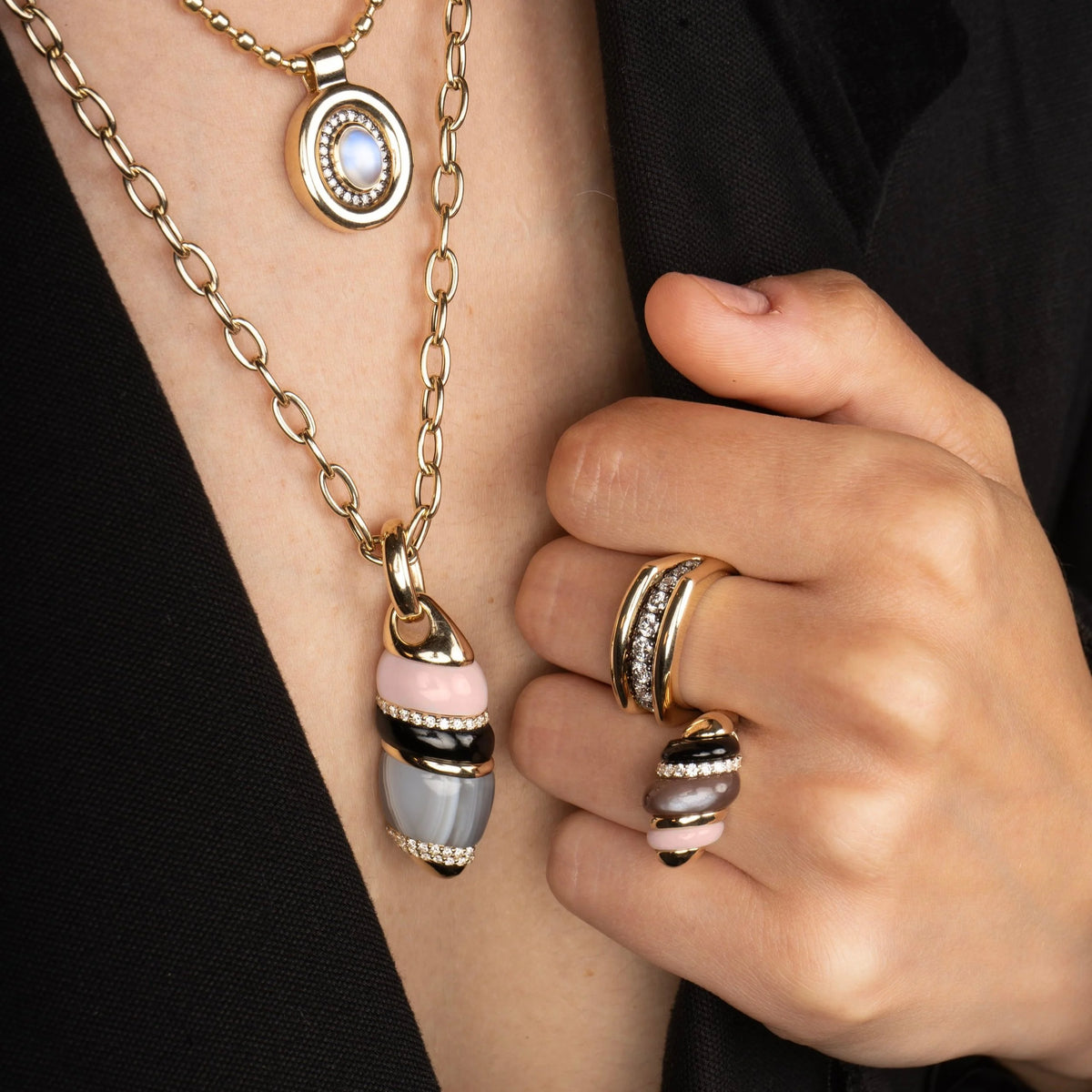 Close-up of a person wearing layered gold-tone jewelry, including a chain necklace with a pendant and stacked rings on the hand, against a black top background.