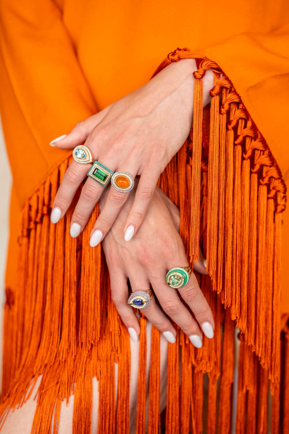 A close-up of a person's hands wearing multiple colorful rings, styled with a bright orange fringed garment.