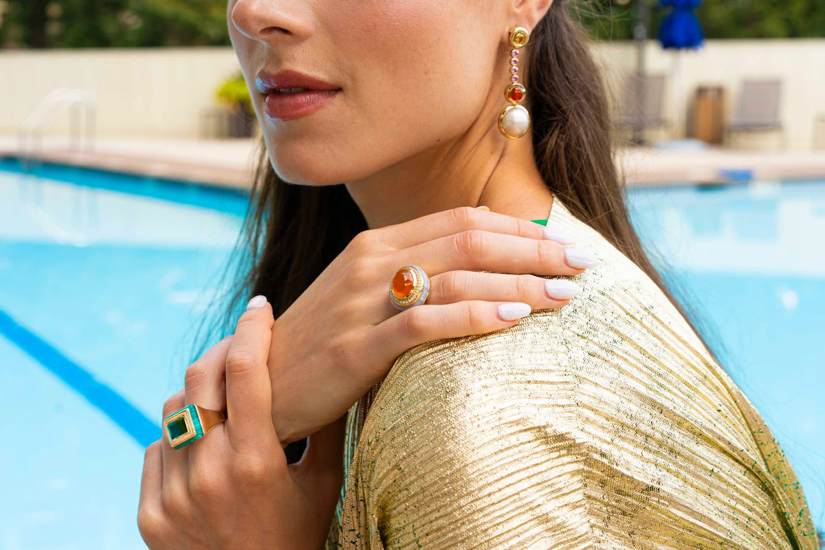 A close-up lifestyle shot of a woman wearing gold jewelry, including a gold ring with a red and white stone and a gold dangle earring with a pearl, set against a blurred swimming pool background.