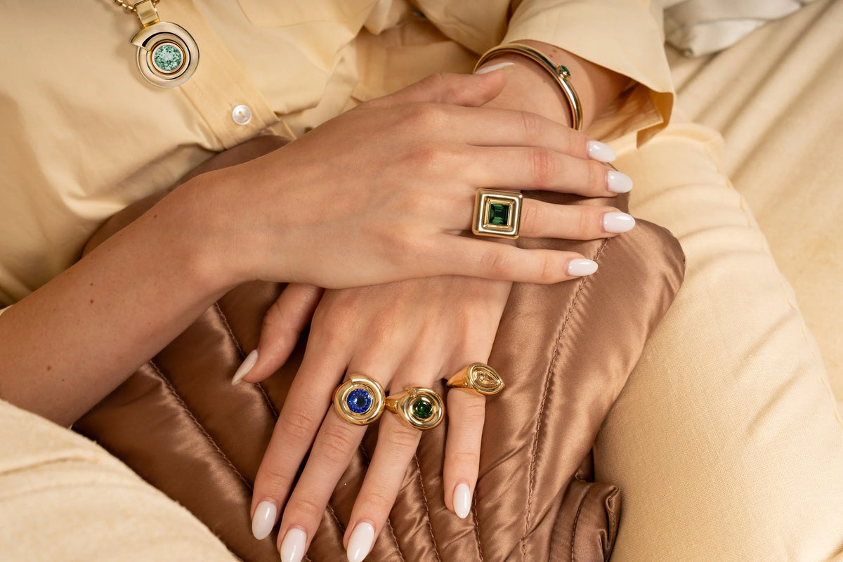A close-up of a person's hands wearing multiple gold rings with green, blue, and oval-cut stones, styled with a beige blouse and a brown satin clutch.