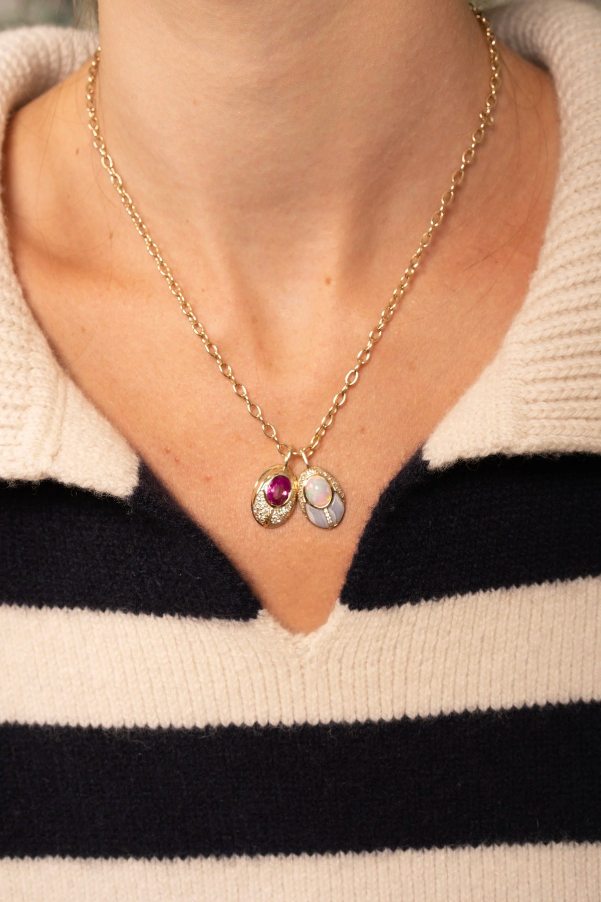 A close-up of a person wearing a delicate gold chain necklace with two small oval pendants, one featuring a pink gemstone and the other with a white inlay, styled over a black and cream striped knit sweater.