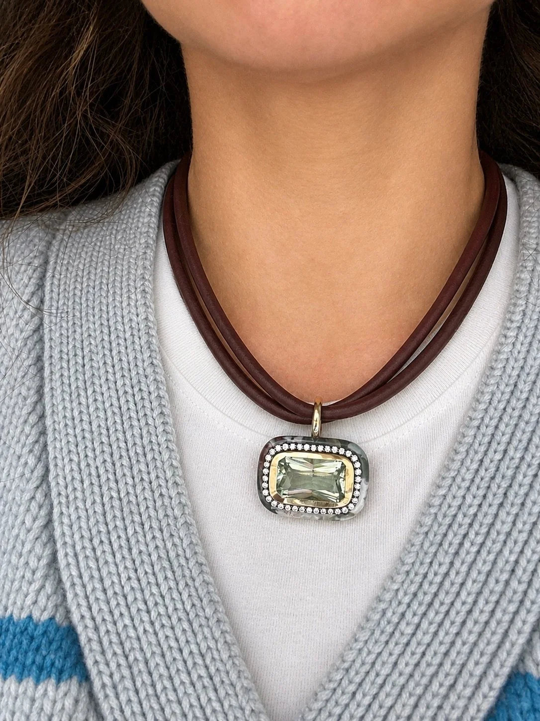 A close-up of a person wearing a multi-strand dark brown leather necklace with a rectangular pendant. The pendant features a light green gemstone surrounded by a silver-tone halo and a border of small clear rhinestones, worn over a white shirt and a gray knit cardigan.