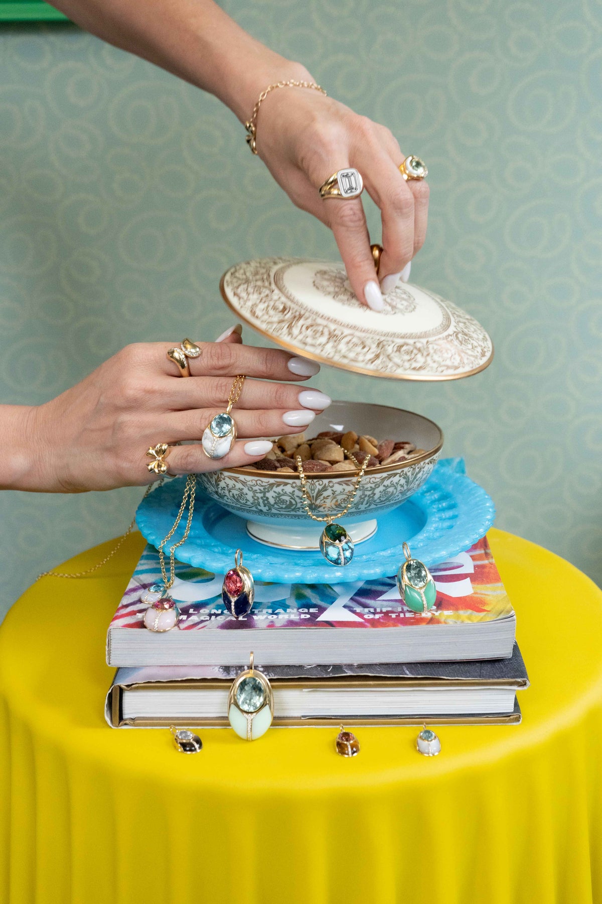 A pair of hands with white manicured nails and gold jewelry opens a decorative ceramic lidded box resting on a stack of books and blue plates. The box contains jewelry, including a blue beaded necklace and a pendant, set against a patterned green wall and a bright yellow table.