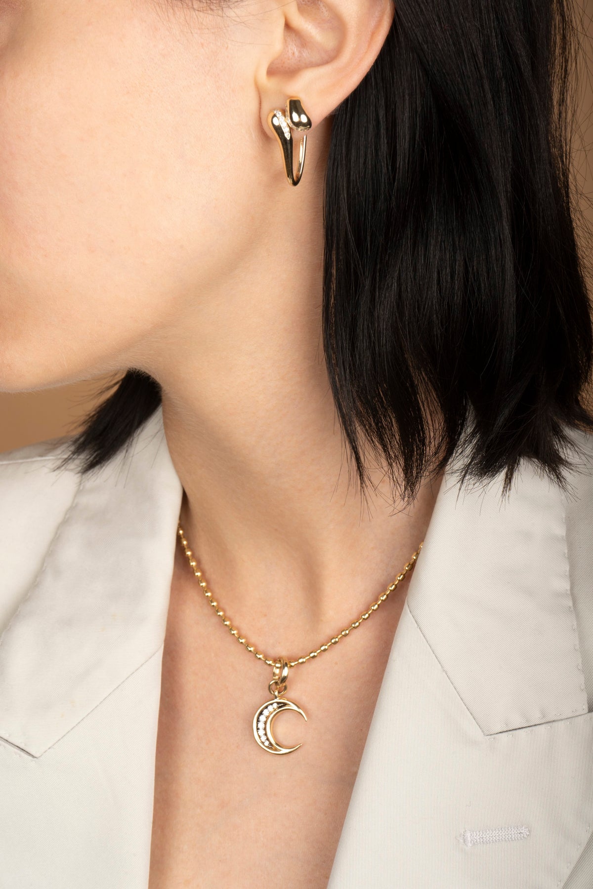 Close-up of a model wearing a gold crescent moon pendant necklace and matching gold hoop earrings, styled with a white blazer.