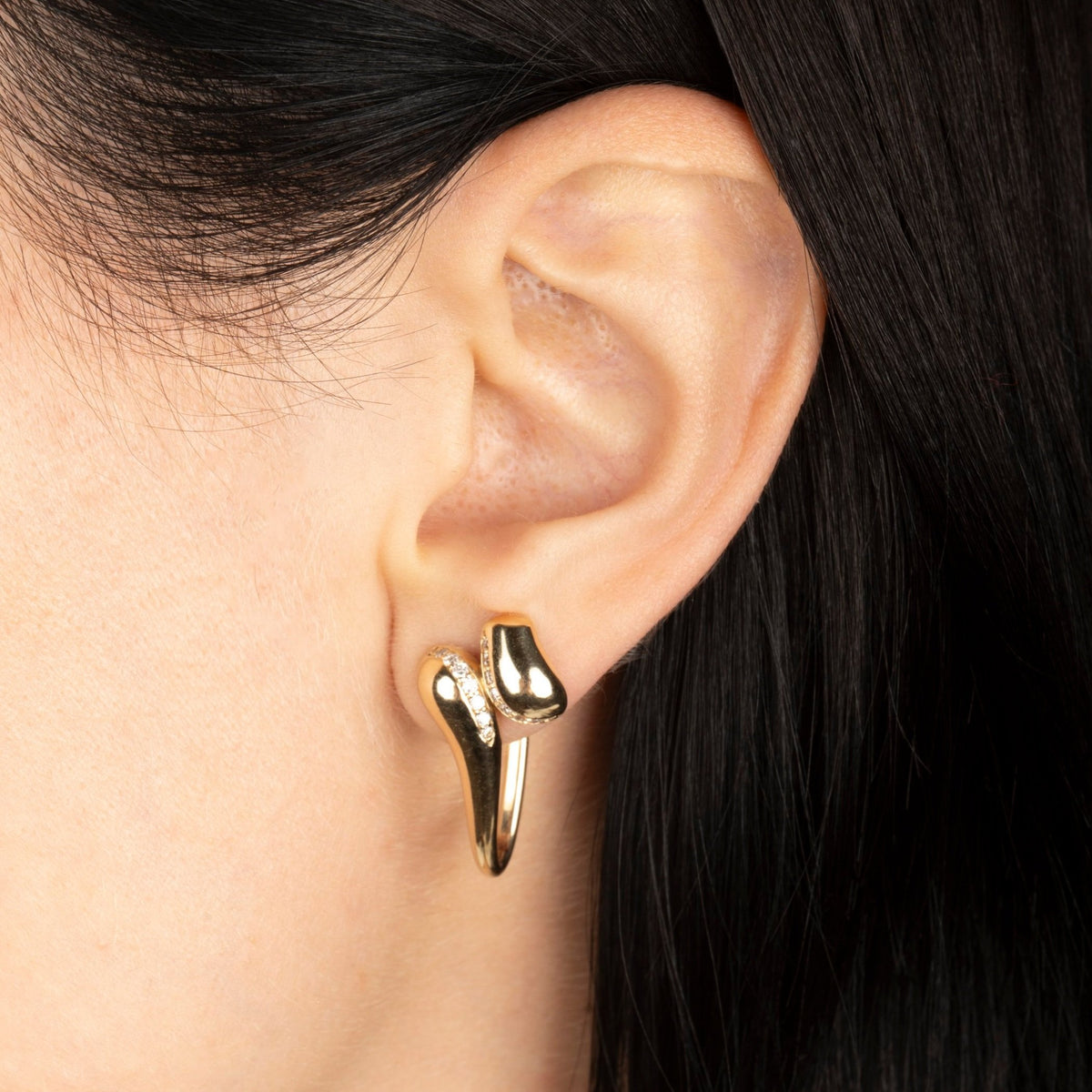 A close-up of a person's ear wearing a polished gold huggie hoop earring with two abstract curved shapes and a small diamond accent.
