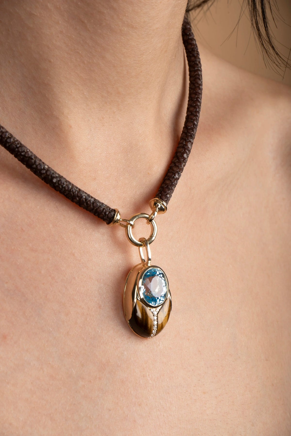 A close-up of a person's neck and chest wearing a dark sting ray leather necklace with a gold-toned pendant featuring a light blue oval gemstone.