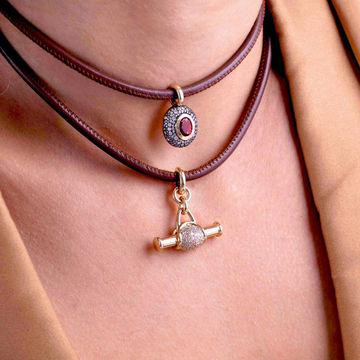 A close-up of a person's neck and chest wearing a double-layered brown leather choker necklace. The top layer features a silver-toned oval pendant with a red gemstone, and the bottom layer features a gold-toned toggle clasp charm.