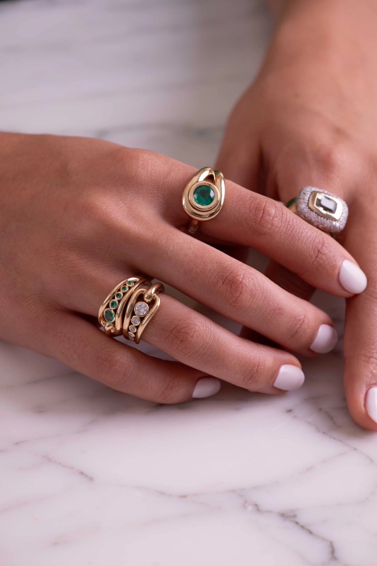 A close-up of a person's hands wearing multiple gold rings with green and white stones, resting on a white marble surface.
