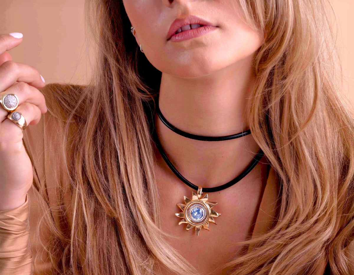 A close-up lifestyle shot of a woman wearing a black choker necklace with a gold sunburst pendant featuring a blue circular center, styled with gold rings.