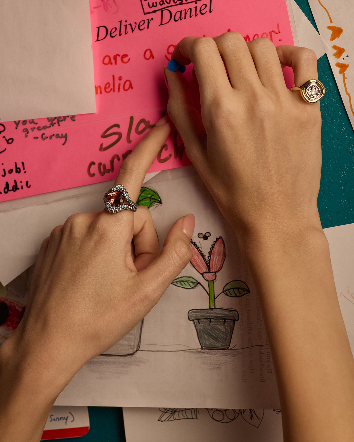 Two hands shown wearing gold rings, interacting with colorful sticky notes and a sketchbook on a desk.