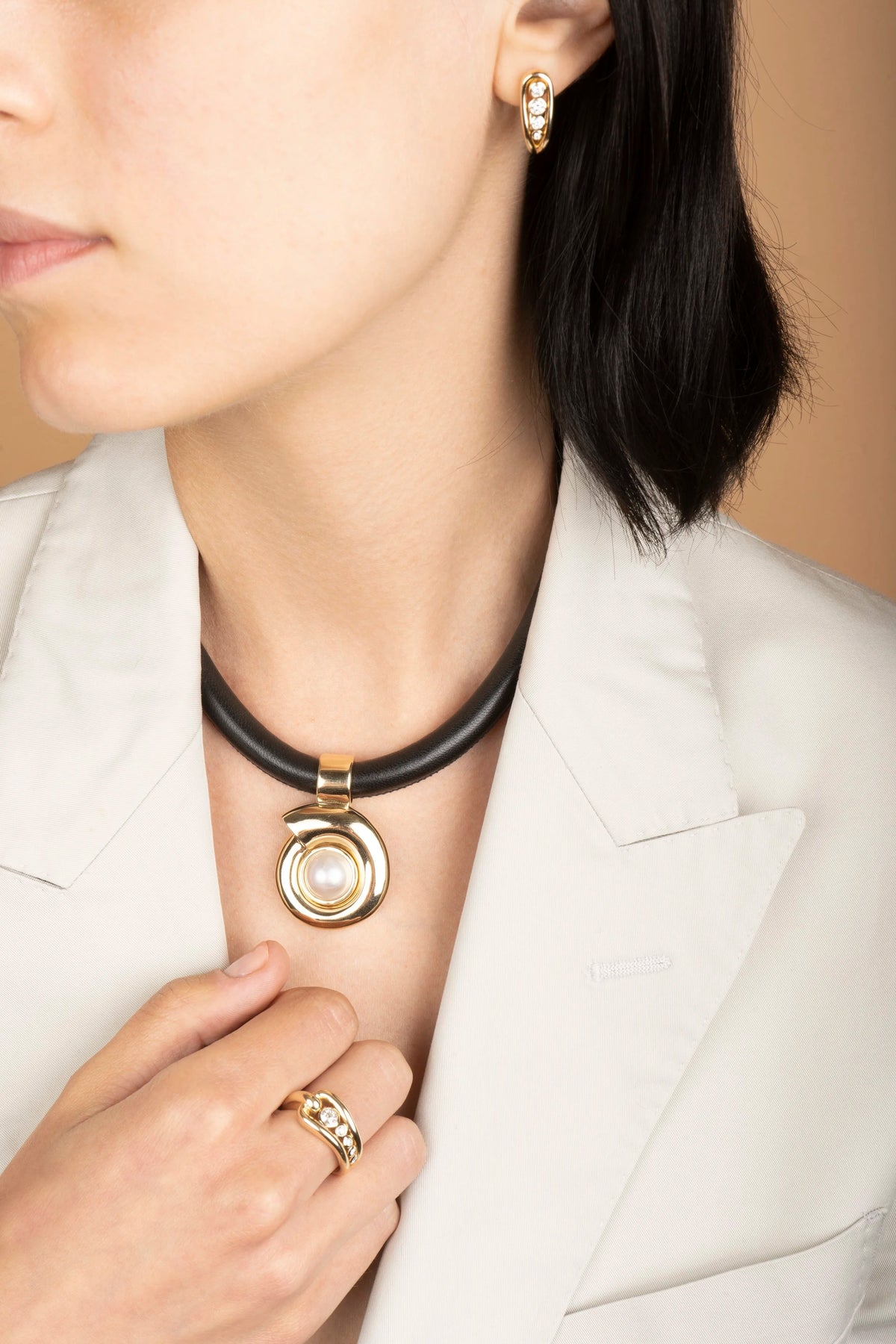 Close-up of a woman wearing a black collar necklace with a gold spiral pendant featuring a white pearl center, paired with matching gold rings and earrings.