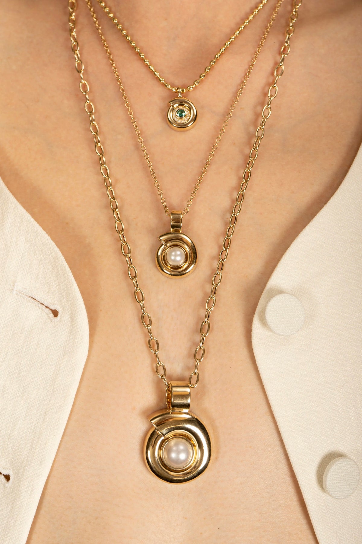 Three layered gold necklaces with circular pendants, including two with white pearls and one with a small green stone, worn on a model's neck over a white collared shirt.