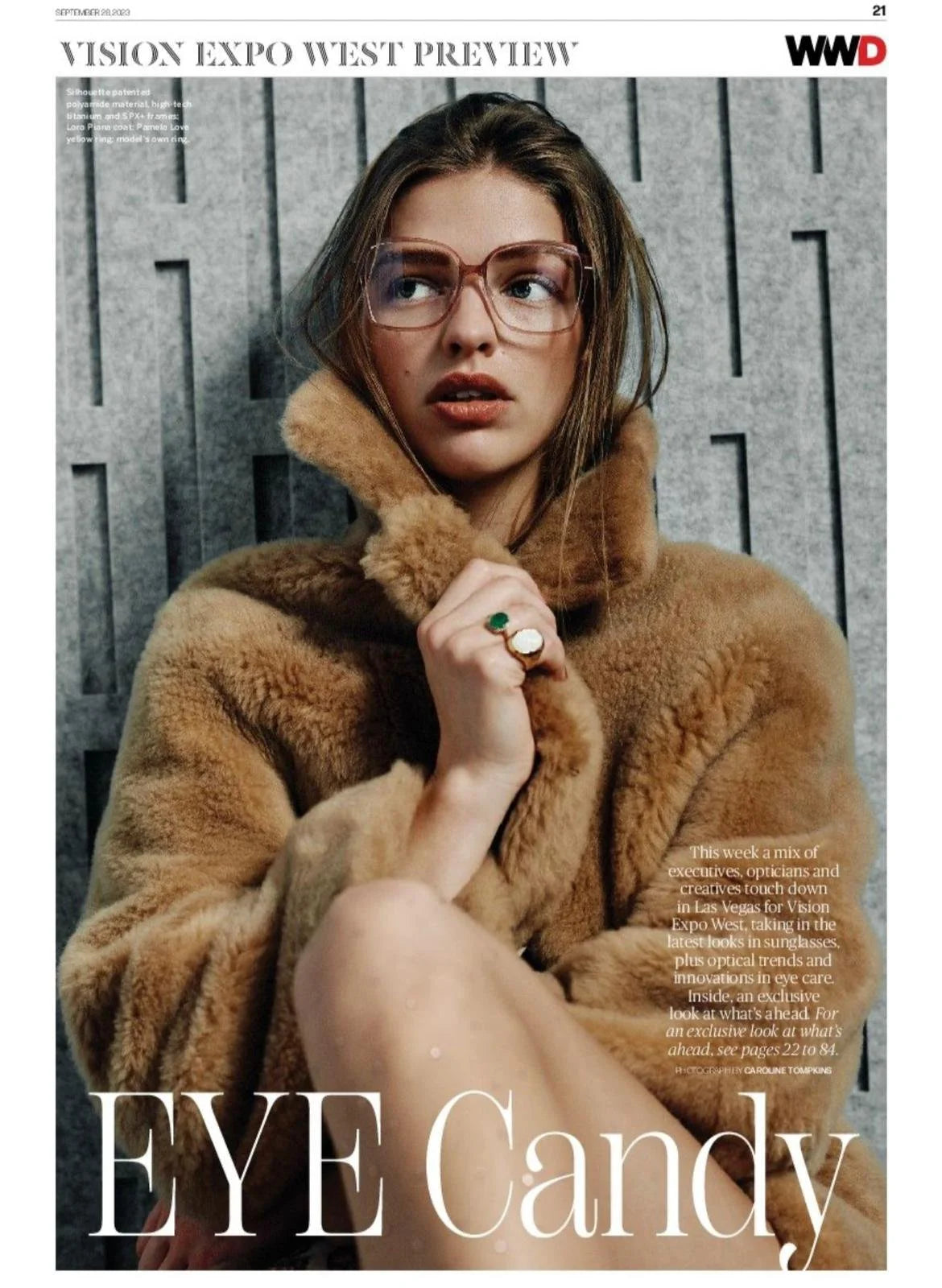 A woman wearing sunglasses and a tan faux fur coat sitting against a gray wooden wall, showing a ring with a large green stone.
