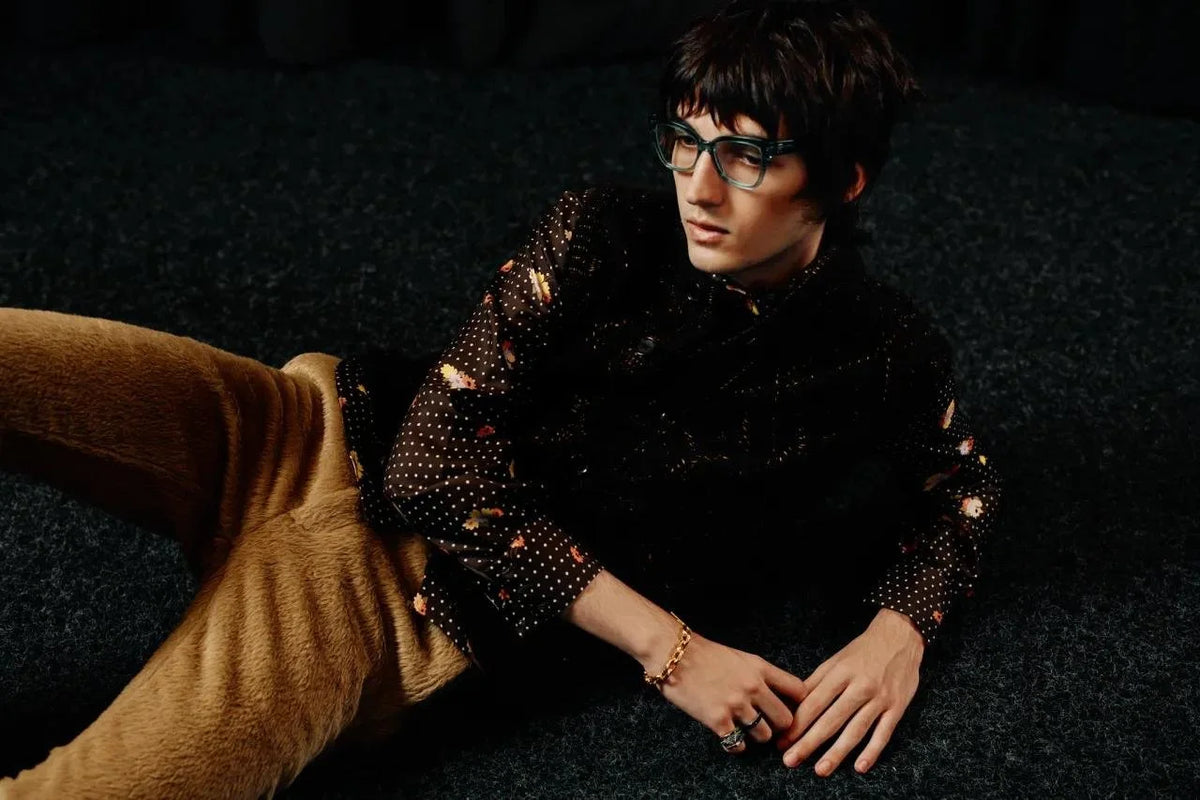 A young man with black hair and glasses, wearing a black shirt with floral and polka dot patterns, is lying on a dark surface, with a large brown dog lying beside him.