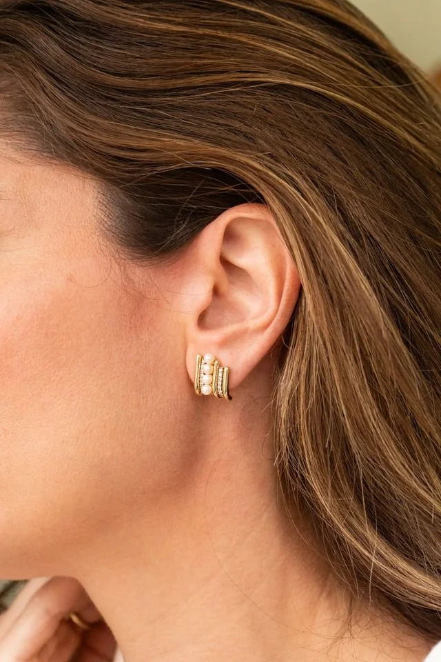 A close-up of a woman's ear wearing a pair of gold huggie hoop earrings with small pearls and diamonds, shown against her brown hair and skin.