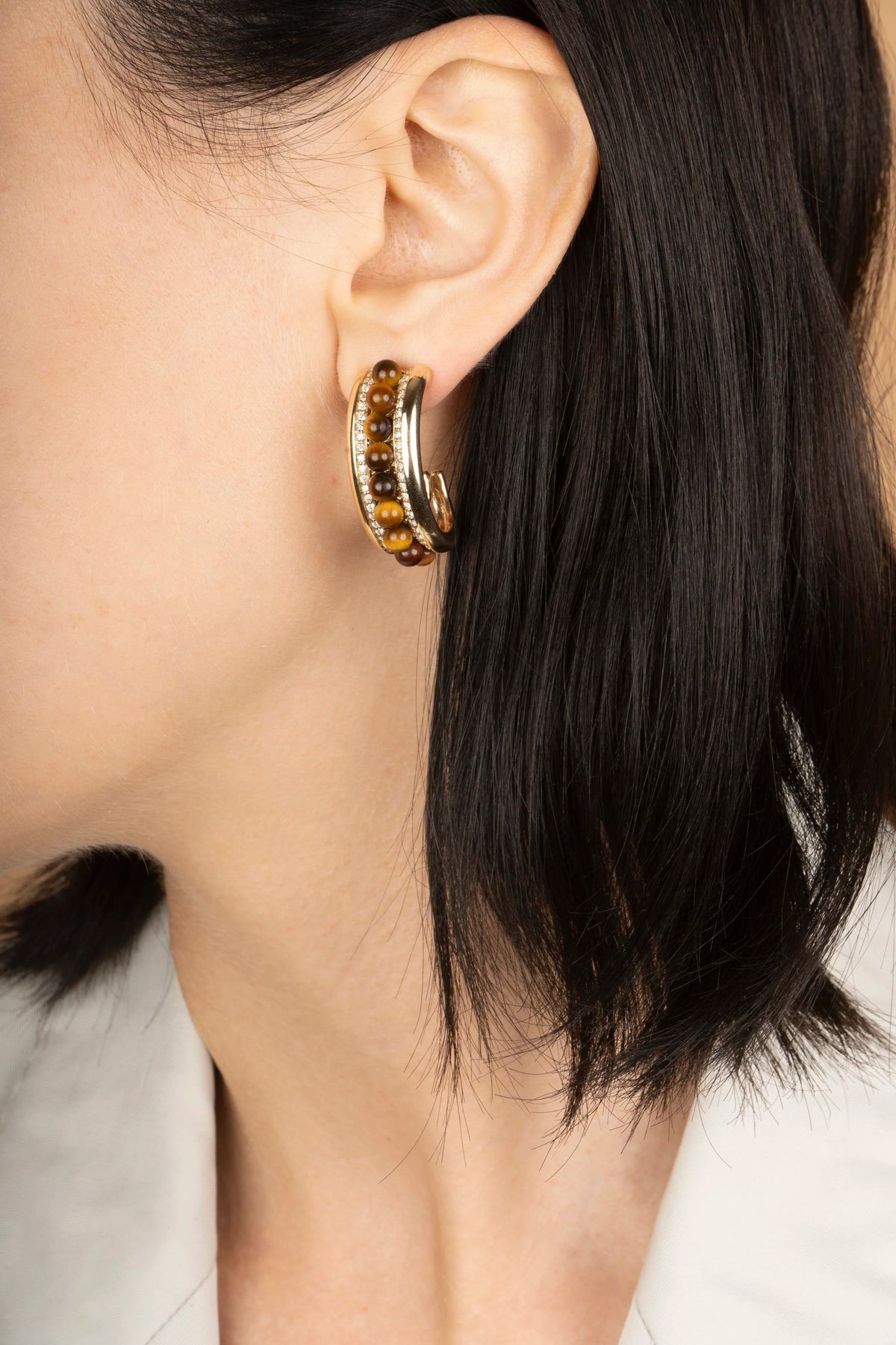 A close-up of a model's ear wearing a gold huggie hoop earring adorned with small amber-colored stones.