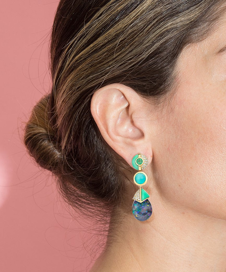 A close-up of a woman's ear wearing a gold drop earring with turquoise and blue gemstone details, set against a solid pink background.