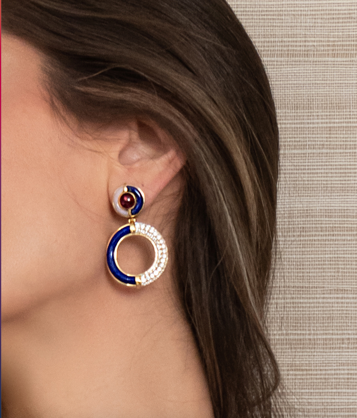 A close-up of a woman's ear wearing a statement drop earring. The earring features a small round stud with a dark red center and a white outer ring, connected to a larger circular drop with a deep blue interior and a white crystal-encrusted outer ring, all set in gold-tone metal.