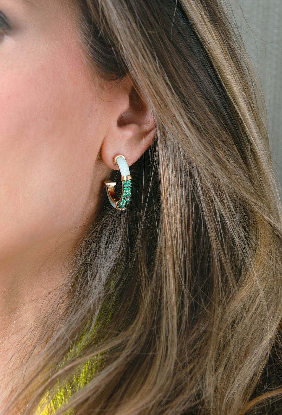 A close-up of a woman's ear wearing a small gold huggie hoop earring with a mint opal top and a curved row of green stones along the front.