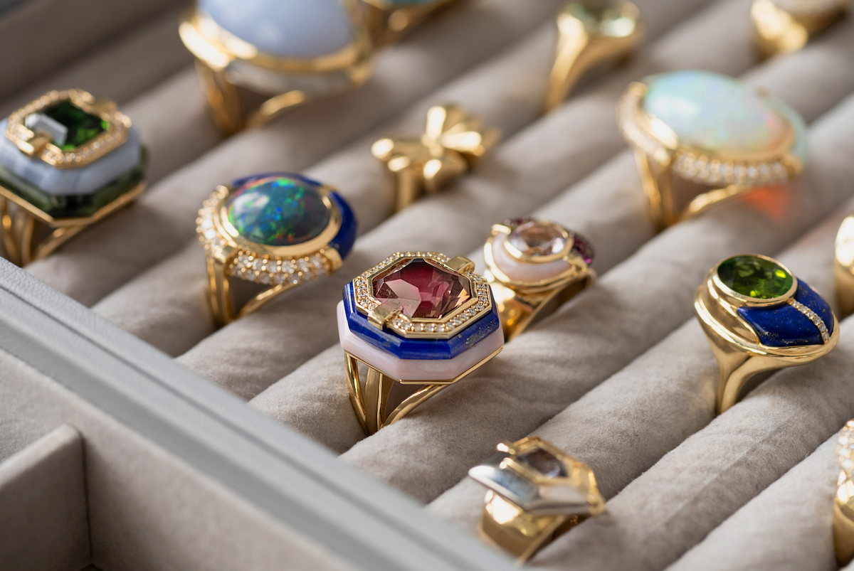 A close-up of multiple gold rings displayed in a gray ring box. The rings feature colorful gemstones including blue, green, pink, and white, with some adorned with small clear crystals.