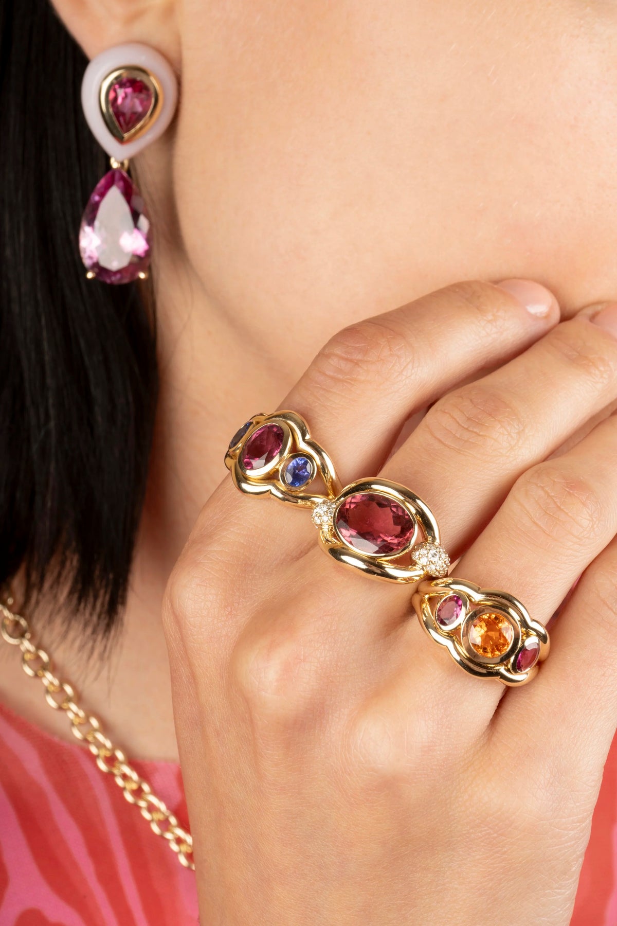 A close-up lifestyle shot of a model wearing a multi-piece gold jewelry set, including a matching drop earring and three rings with large pinkish-red, blue, and orange gemstones.
