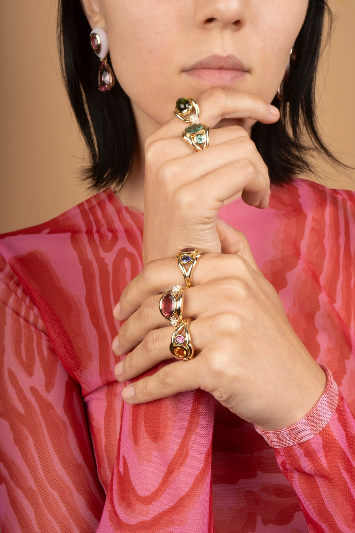 A close-up lifestyle shot of a model wearing multiple gold rings with colorful gemstones on her fingers, paired with a pink patterned top against a warm beige background.