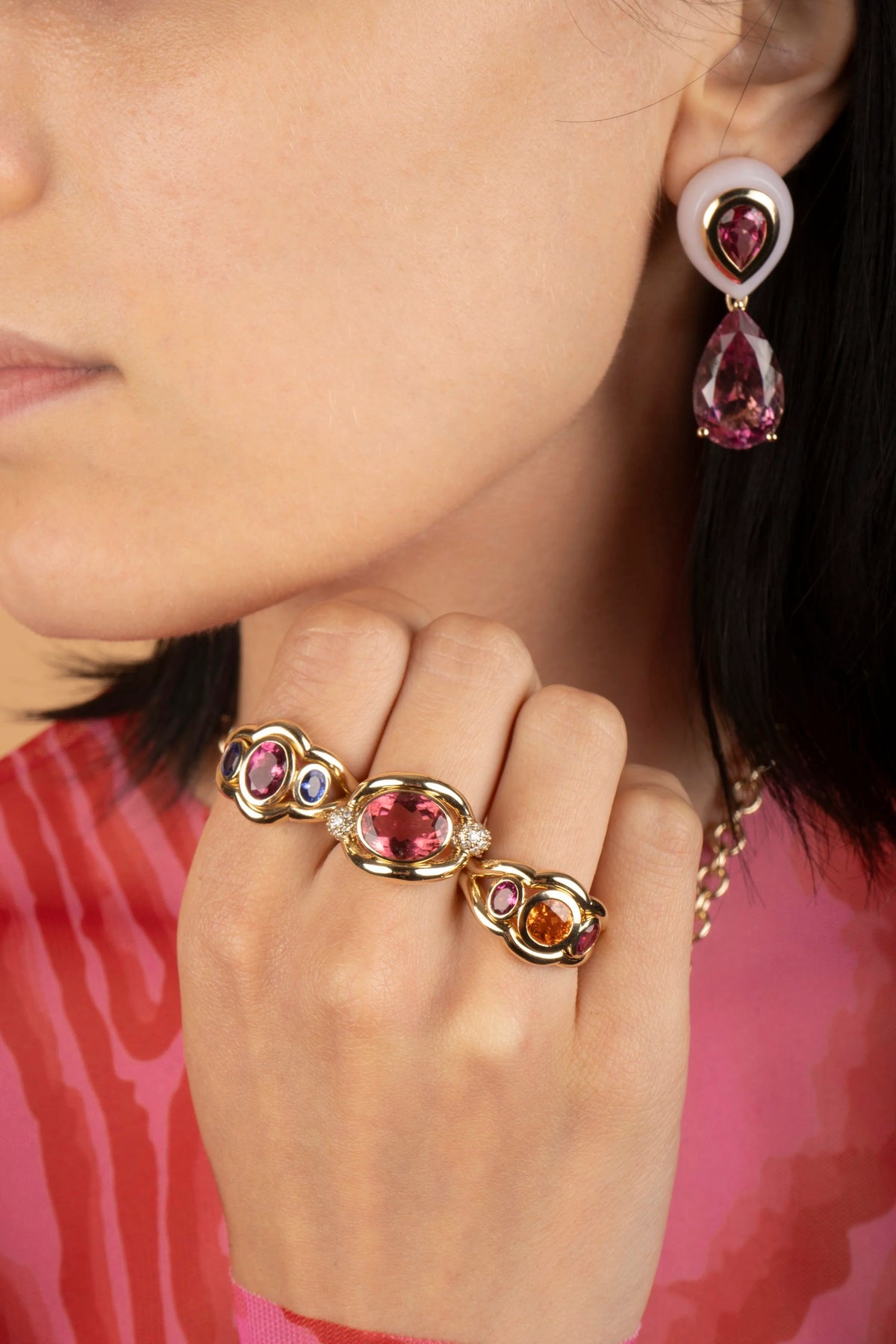 A close-up lifestyle shot of a model wearing gold rings with pink, purple, and orange gemstones, along with a matching drop earring.