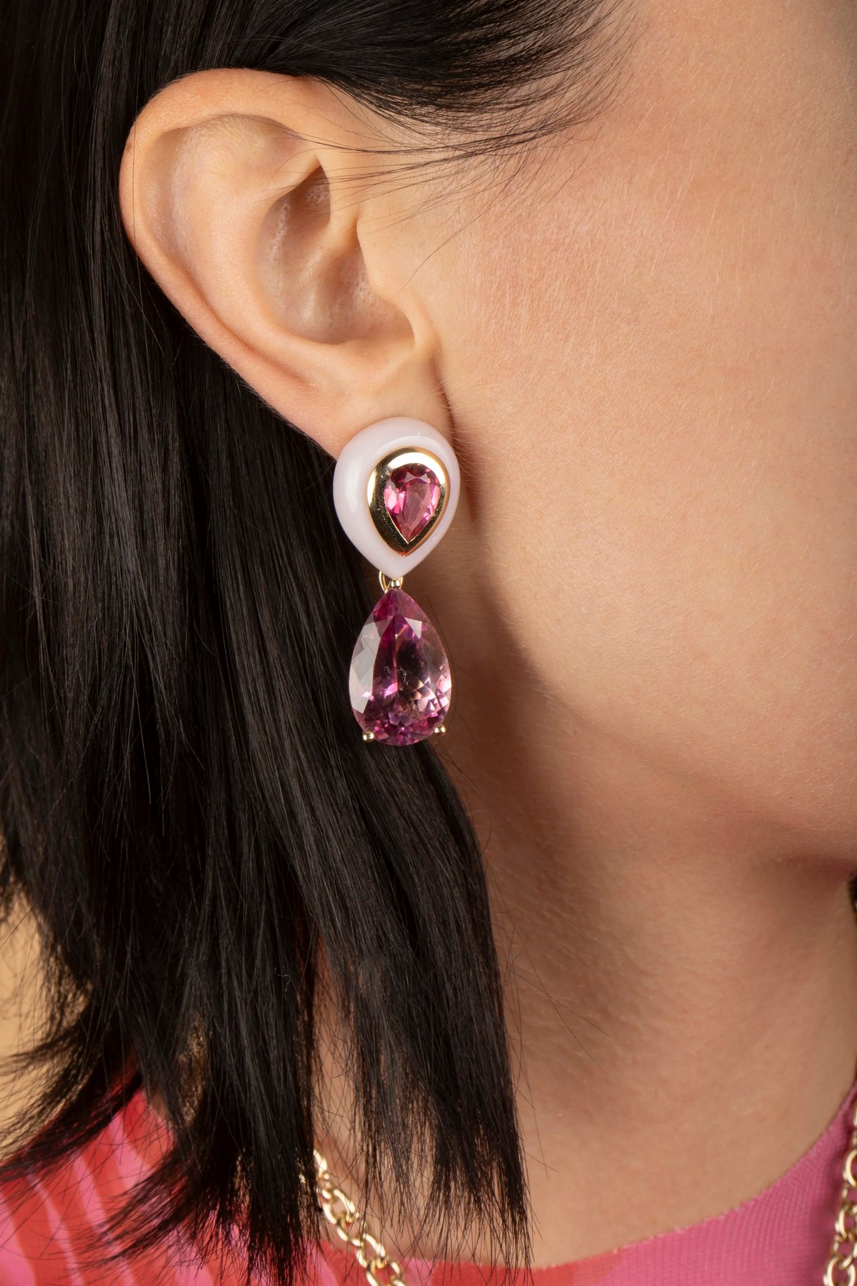 A close-up of a model's ear wearing a dangling earring with a white circular top, a gold heart-shaped setting, and a teardrop-shaped pink gemstone.