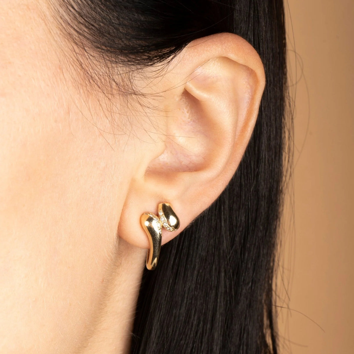 A close-up of a person's ear wearing a small gold huggie hoop earring with a polished, organic curved design.