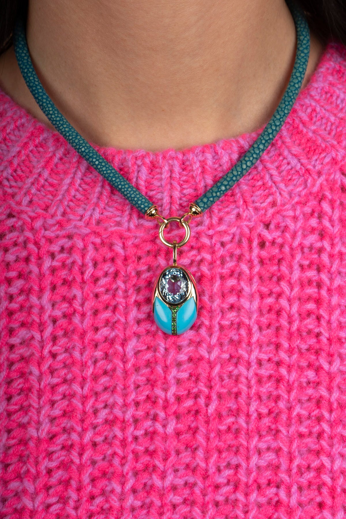 A close-up of a person wearing a turquoise sting ray leather necklace and gold pendant over a bright pink knitted sweater.