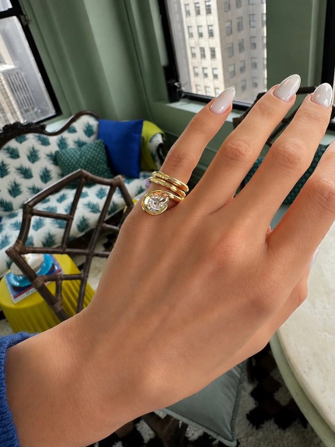 A close-up of a hand wearing a gold ring with a large oval clear gemstone, set against an indoor background with a window and furniture.