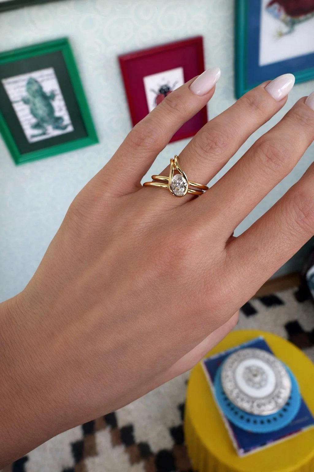 A gold ring with a teardrop-shaped clear gemstone and twisted band details, shown worn on a person's finger. The background features a light-colored wall with framed pictures and a yellow side table.